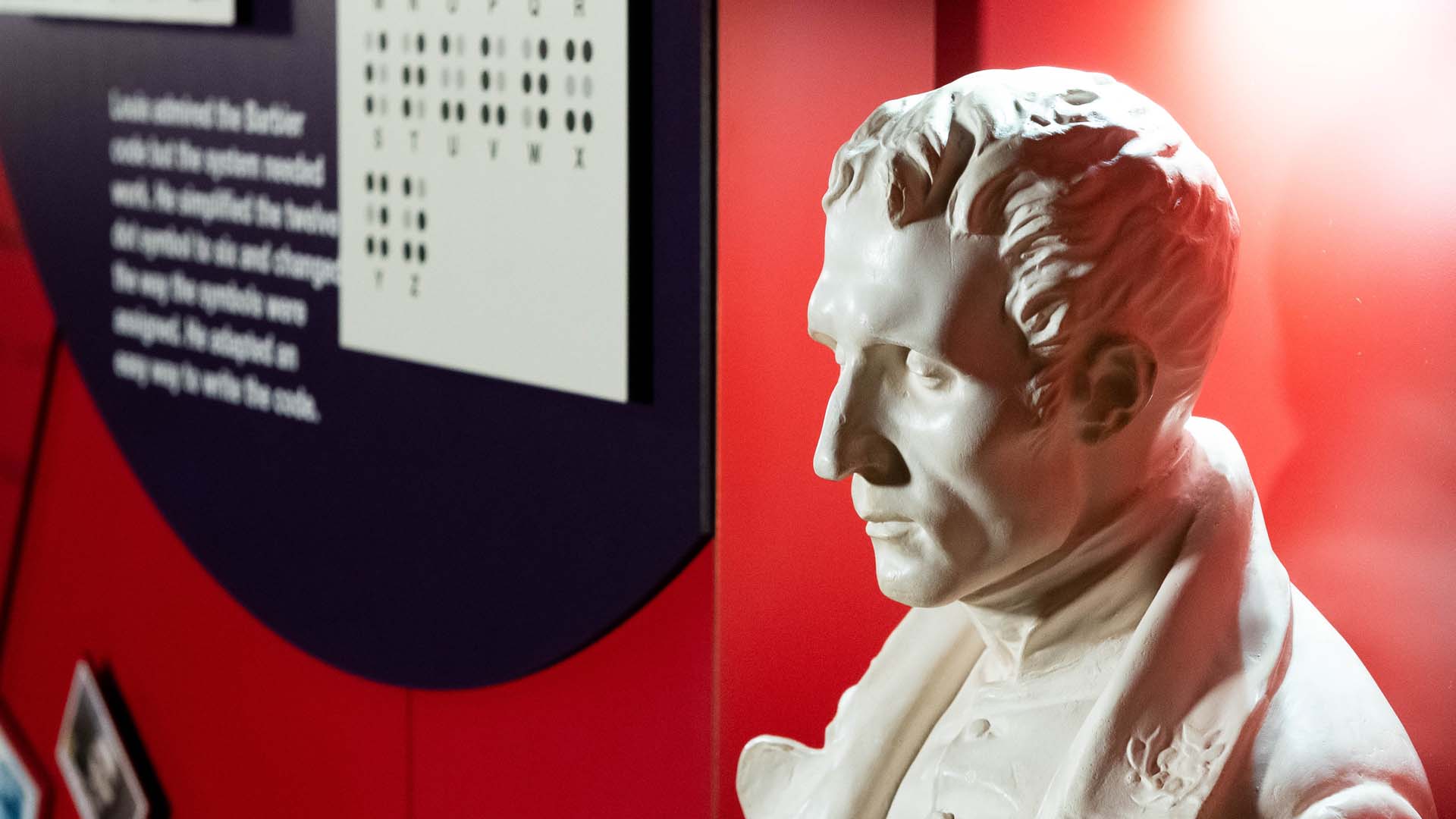 A white plaster bust of Louis Braille beside a museum exhibit wall explaining the braille system