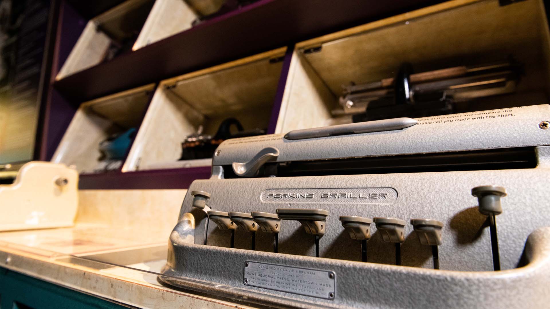 Closeup view of the keyboard of a Perkins Brailler in front of a case full of other historic braillewriters
