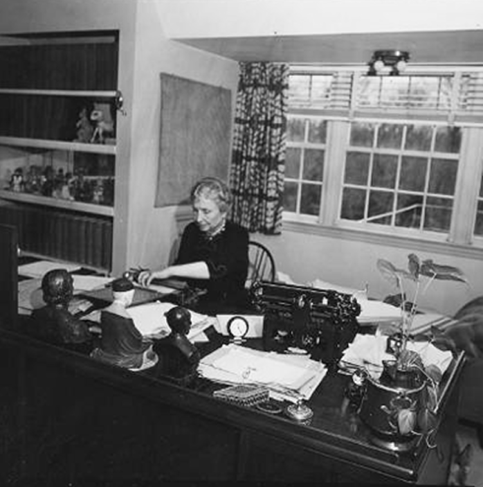 A black-and-white photograph of Helen Keller typing on a braillewriter at a large desk covered with potted plants, stacks of paper, and scattered art objects and paper weights