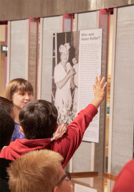 A woman describes a museum exhibit while children crowd in, with one boy raising his hand to get the woman's attention