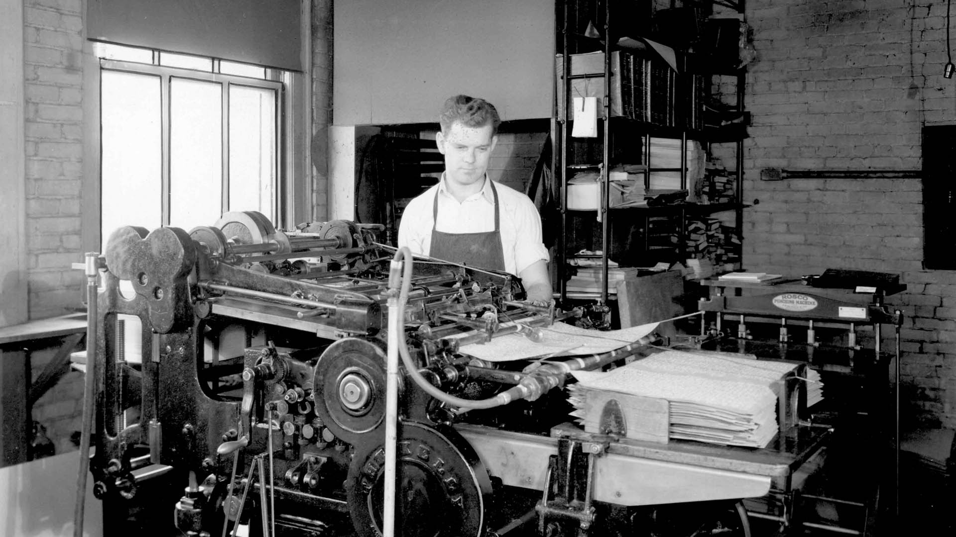 A man wearing an apron stands behind a bulky printing press watching braille pages stack up on the right in a printshop