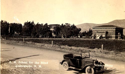 Postcard featuring an antique car parked on a gravel road. In the background are buildings on the campus of the New Mexico School for the Blind.-2