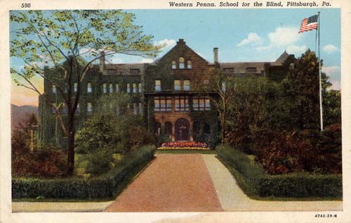 Colored postcard with a large school building. A wide walkway defined by trimmed hedges and trees leads up to flower beds in front of an arched entrance. An American flag blows in the breeze on a flagpole.-1