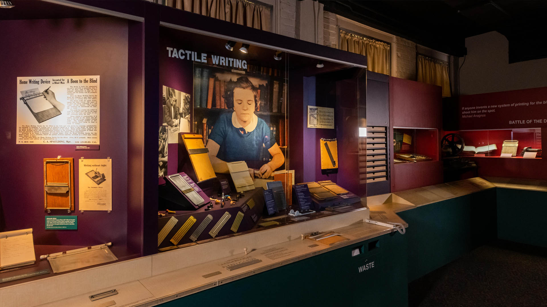 A museum exhibit featuring a case full of braille slates, a picture of a girl writing braille, and a reading rail with tools mounted to it